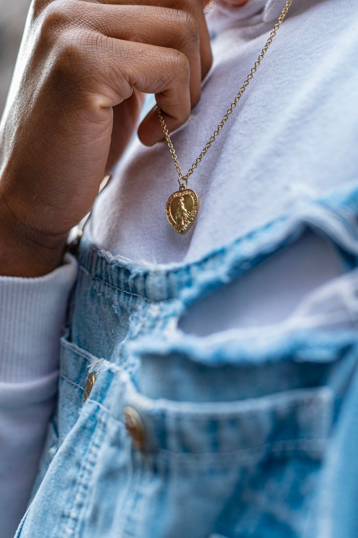 A person holding a gold necklace wearing a white sweater and denim overalls.
